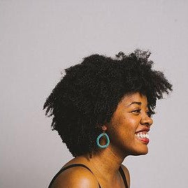 Smiling woman with curly hair and teal hoop earrings against plain gray background